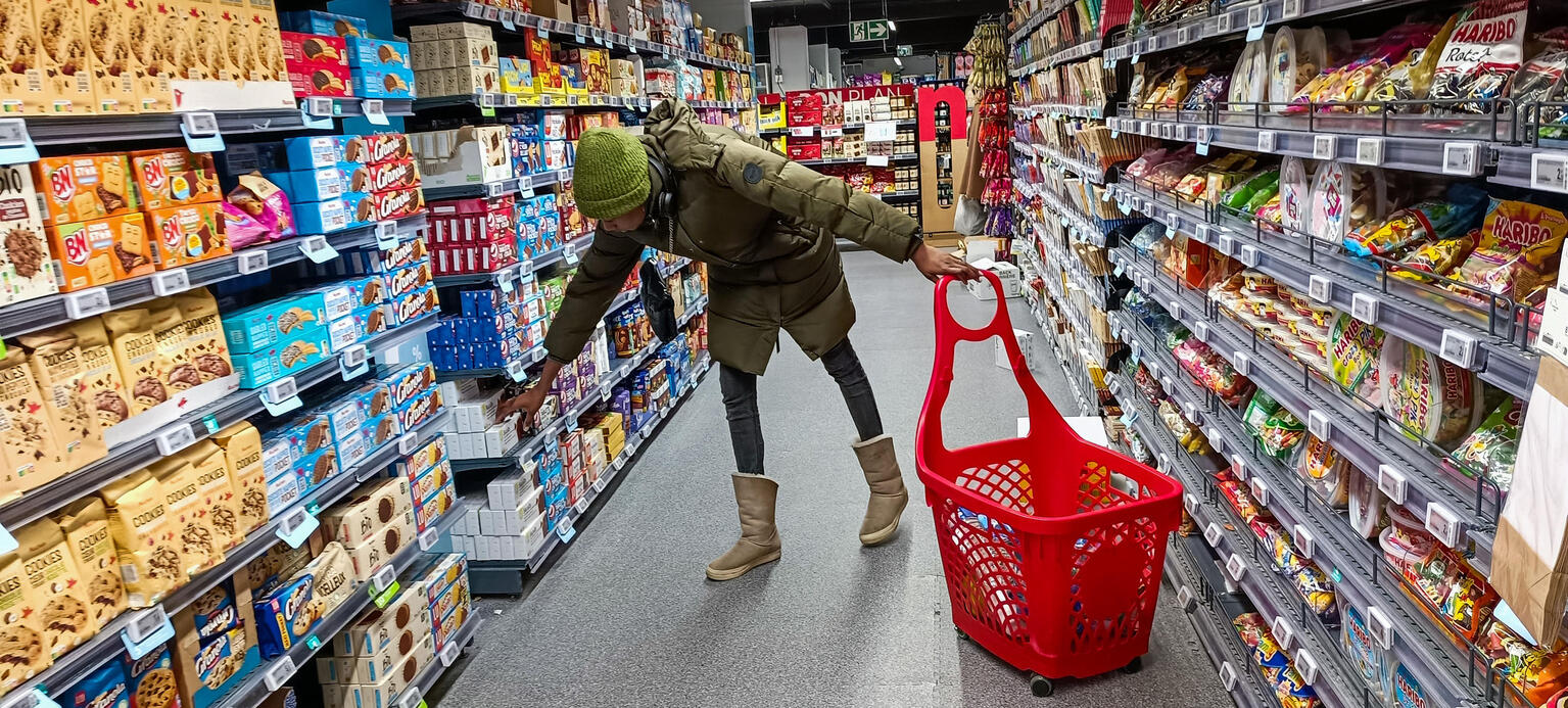 In einem Supermarkt mit vollen Regalen bückt sich eine Frau mit Einkaufskorb um ein Produkt aus dem Regal zu nehmen. Sie hat Winterkleidung an.