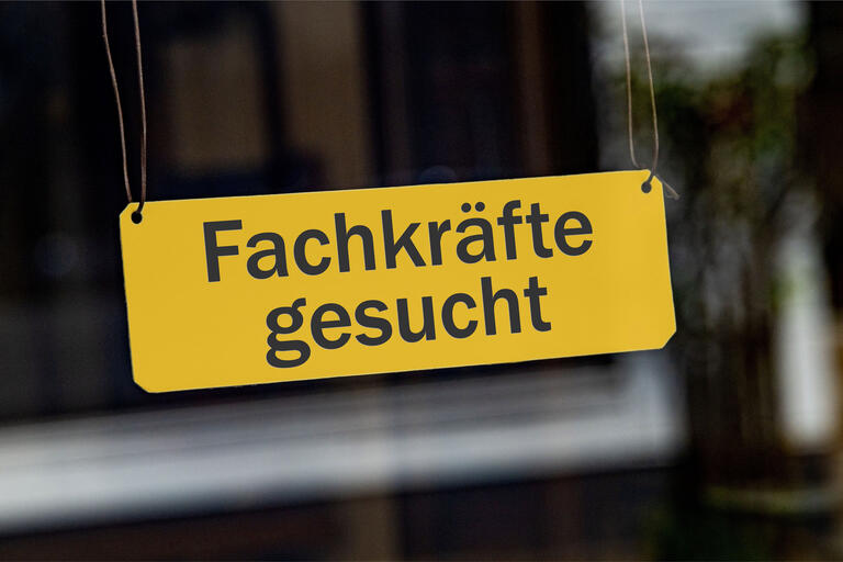 Ein symbolisches, gelbes Schild mit der Aufschrift 'Fachkraefte gesucht' haengt leicht schraeg an Lederbaendern in einem Schaufenster. 