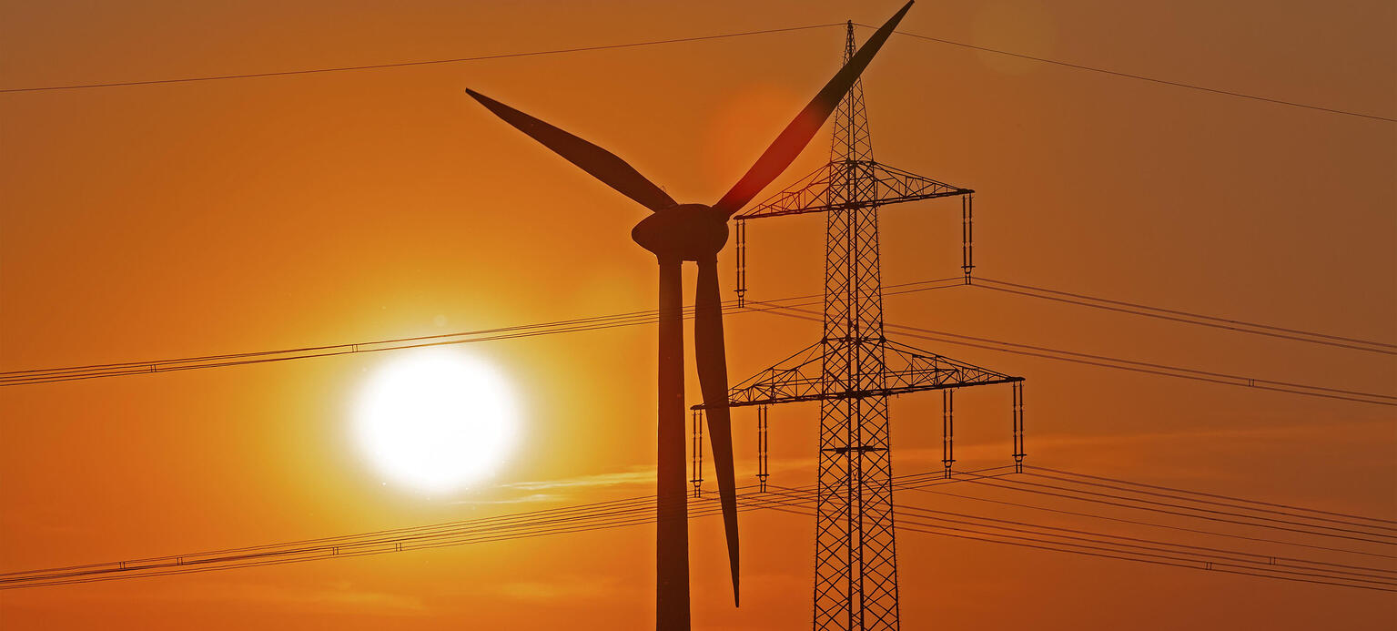 Im Hintergrund geht die Sonne unter und es ist ein orangener Himmel zu sehen. Im Vordergrund sind ein Windkraftwerk und Hochspannungsmast.