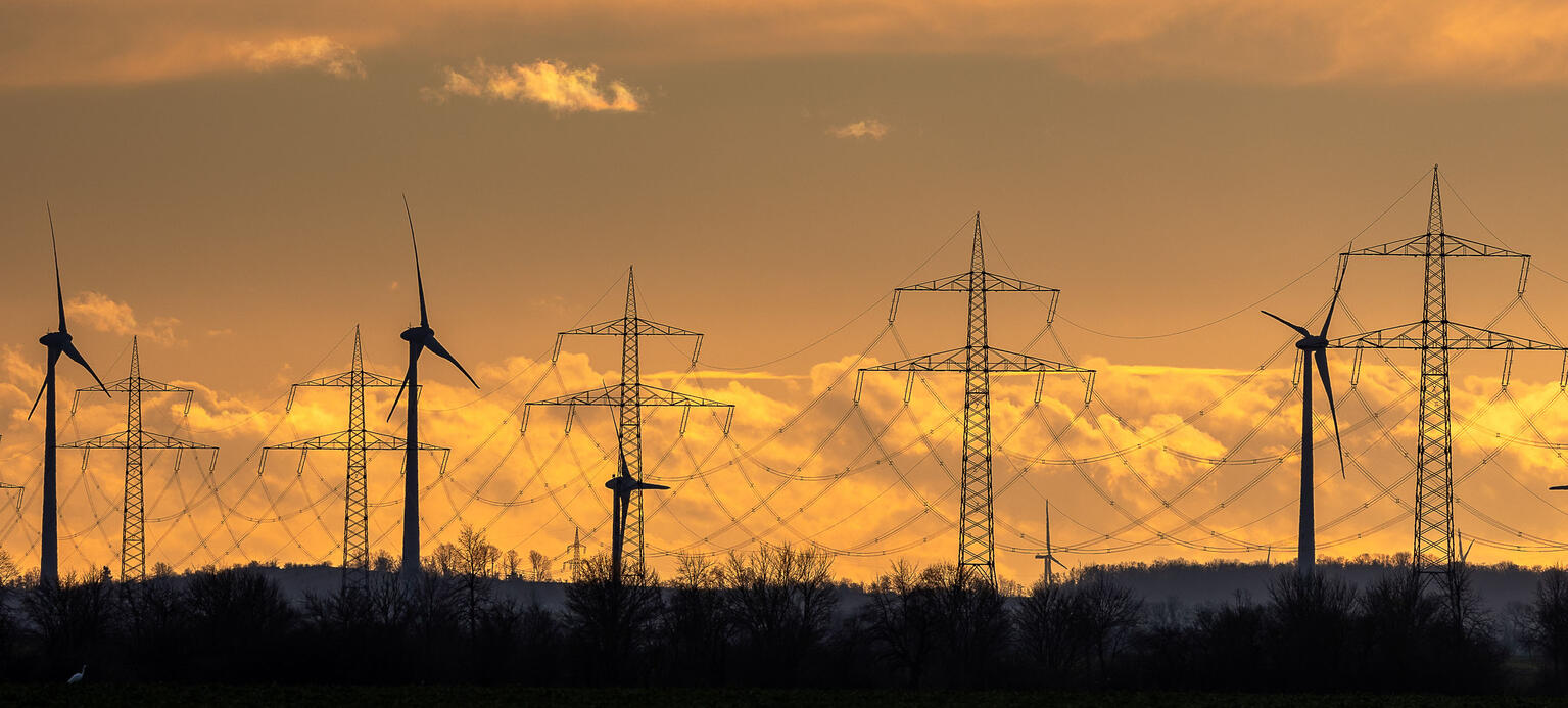 Mehrere Windkraftanlagen und Strommasten stehen vor einem intensiv orangefarbenen Himmel, der durch die untergehende Sonne beleuchtet wird. 