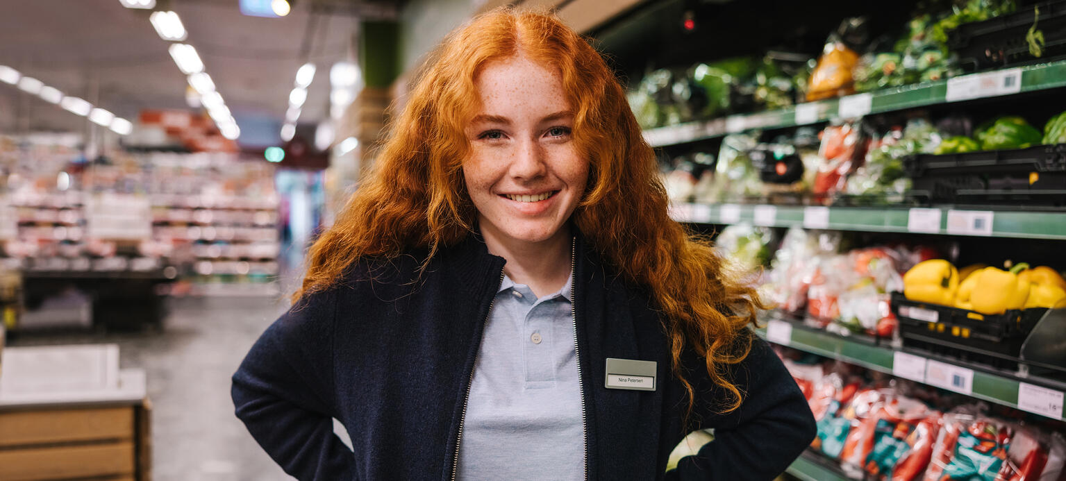 Lächelnde junge Frau (Azubi) steht im Supermarkt vor Obstregal